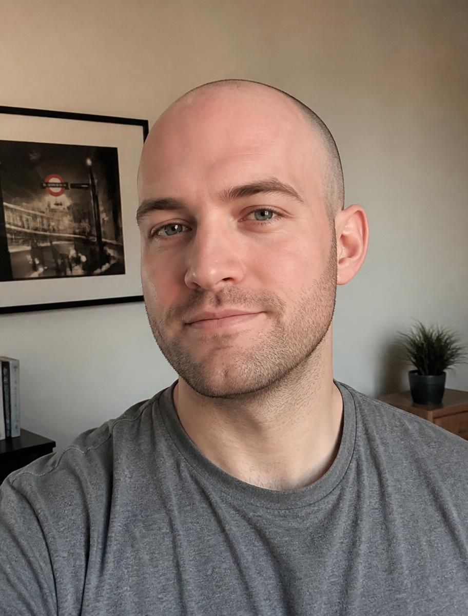 A man in his late twenties with a freshly shaved head and light stubble, at home in a grey t-shirt